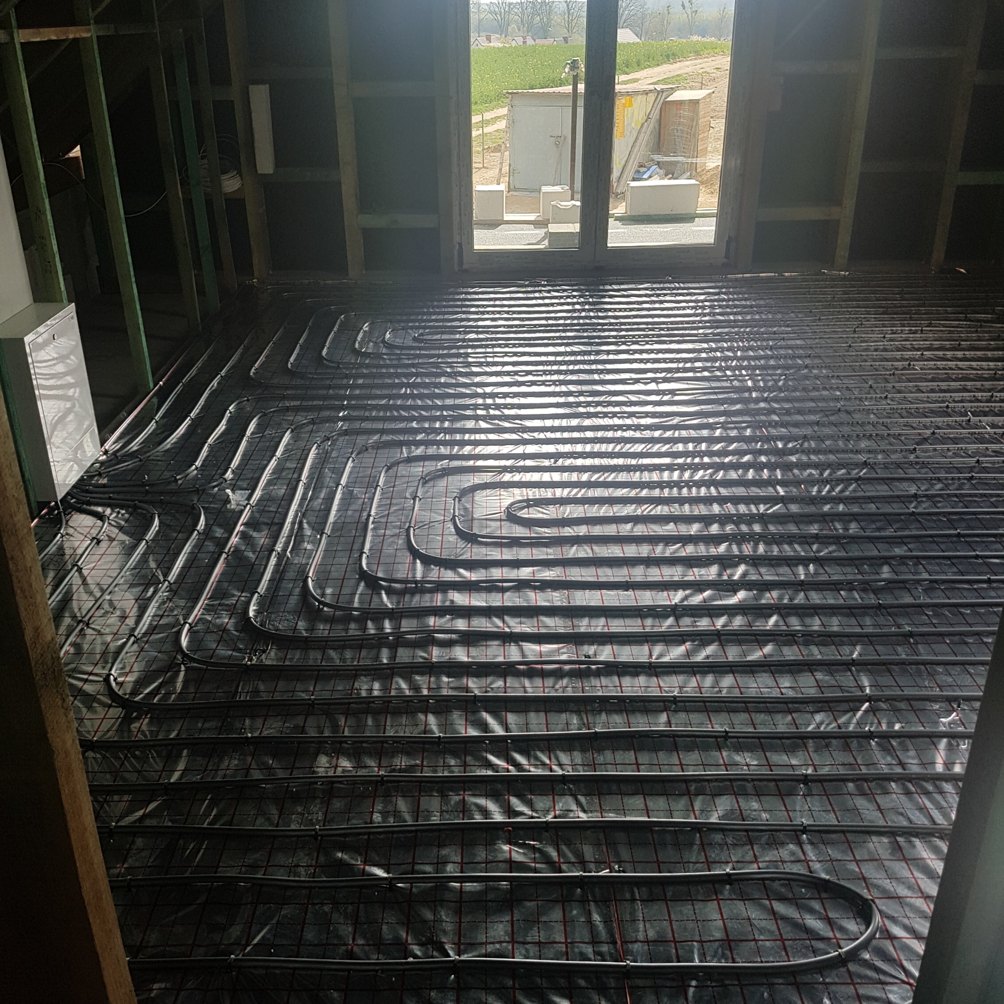 Rozłożona instalacja ogrzewania podłogowego na folii termoizolacyjnej w surowym wnętrzu, widok na okno z zielenią w tle. Elementy konstrukcji budynku.