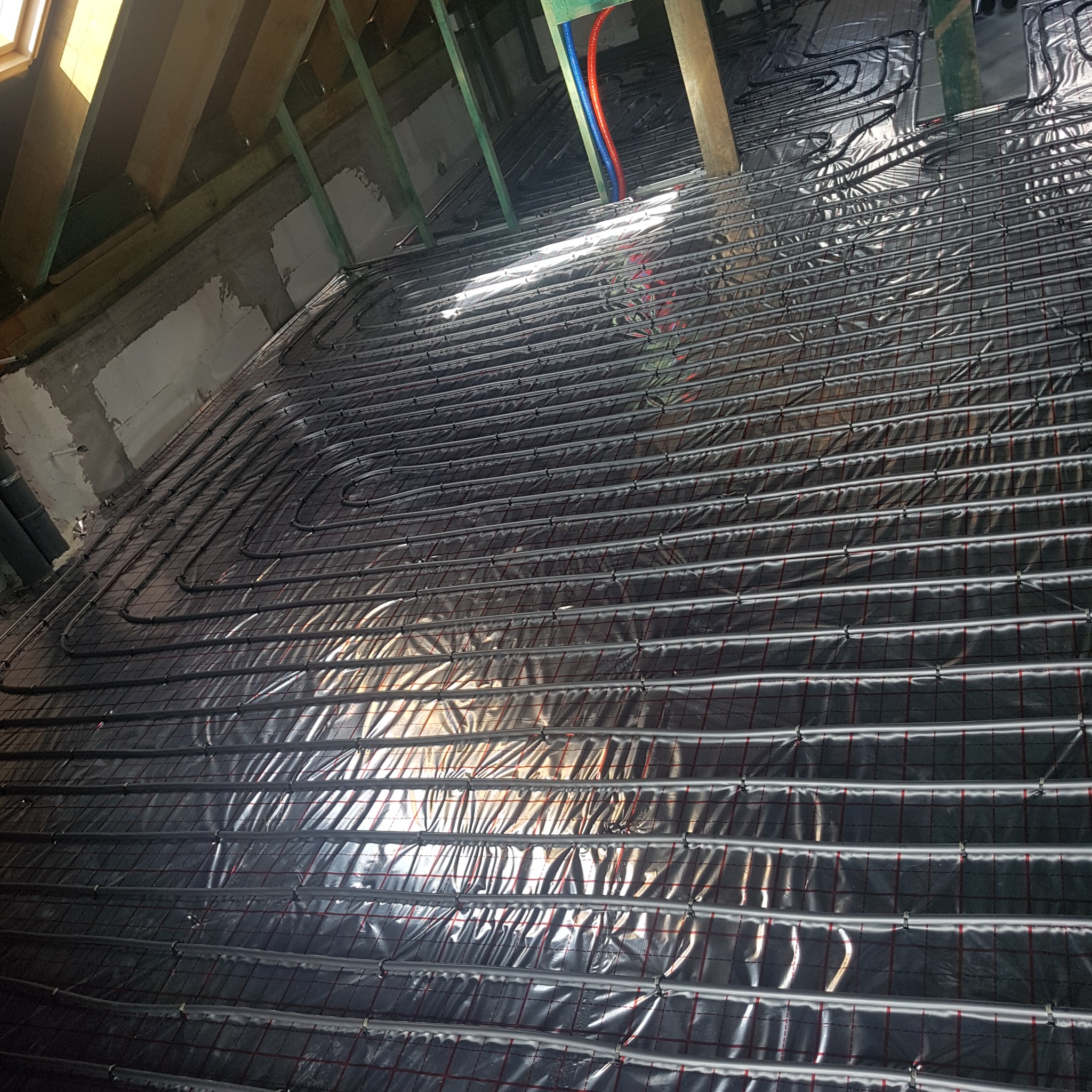 Ułożona instalacja ogrzewania podłogowego na folii w Prabutach. Widoczne rury grzewcze na izolacji, przygotowane do zalania wylewką. Elementy konstrukcji dachu w tle.