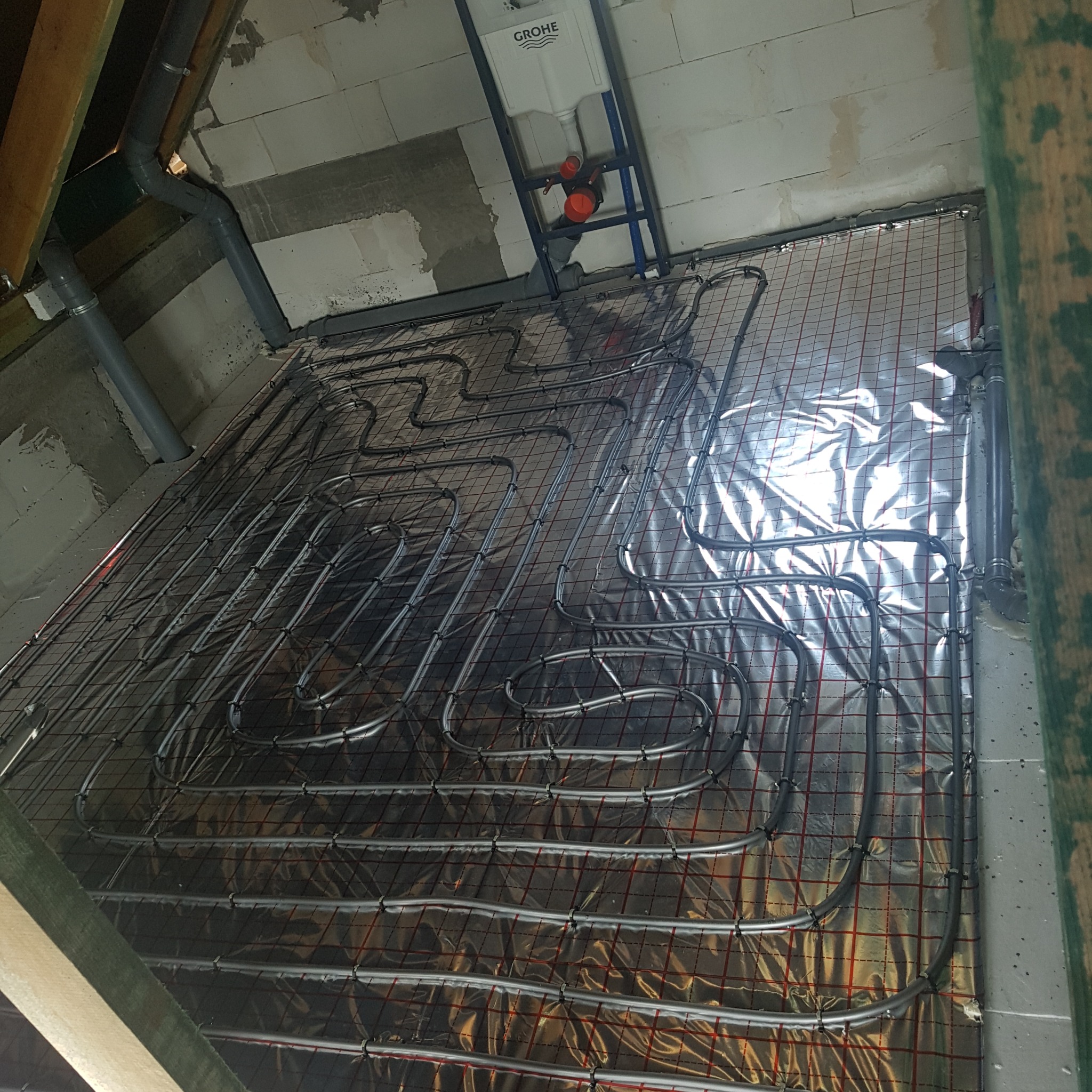 Instalacja ogrzewania podłogowego na folii z siatką, rury układane w pętle, widoczne elementy instalacji sanitarnej Grohe i rury odpływowe. Etap prac przed zalaniem.