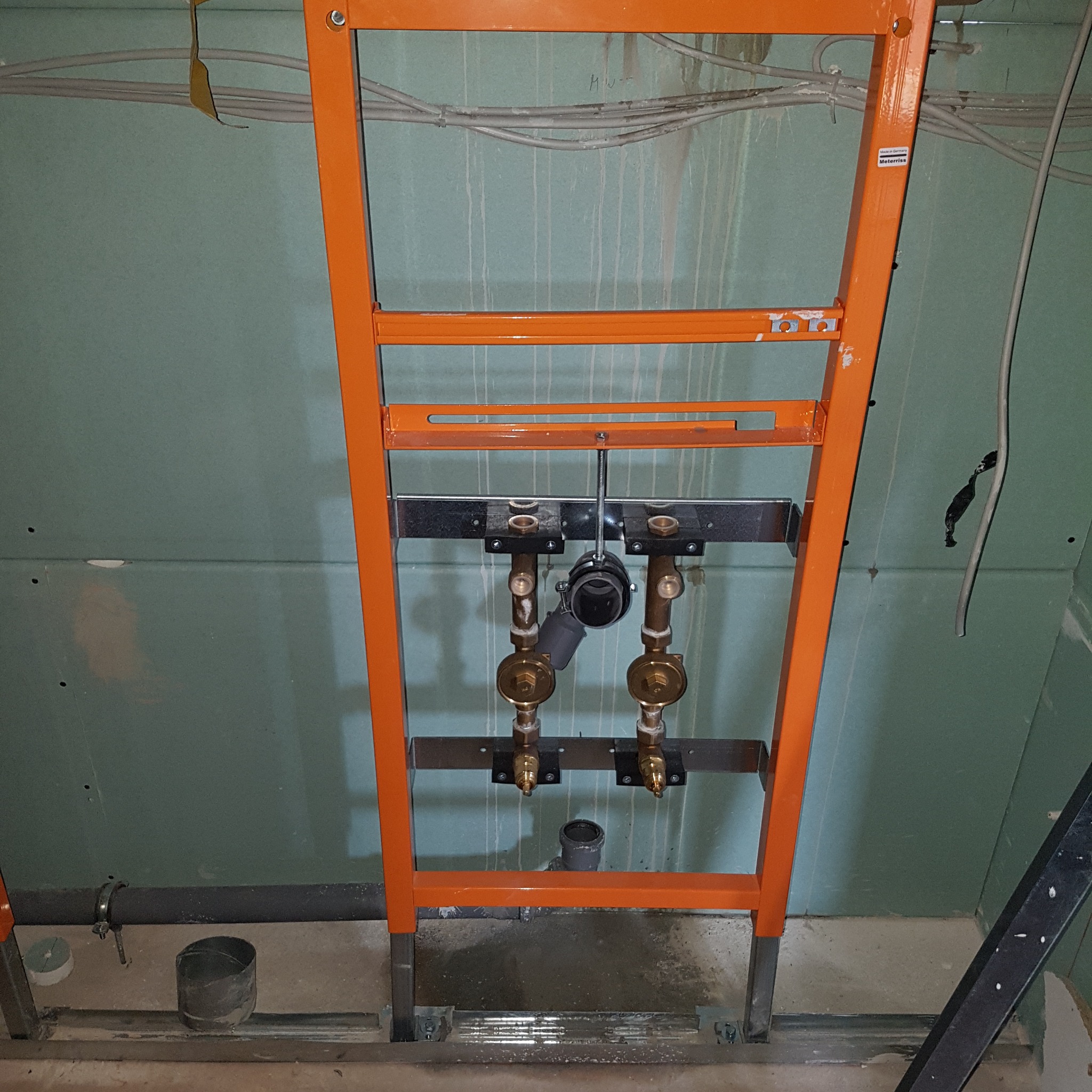 Pomarańczowa rama montażowa WC na tle zielonej płyty gipsowej, widoczne przyłącza wodne i kanalizacyjne. Elementy instalacji w trakcie montażu.