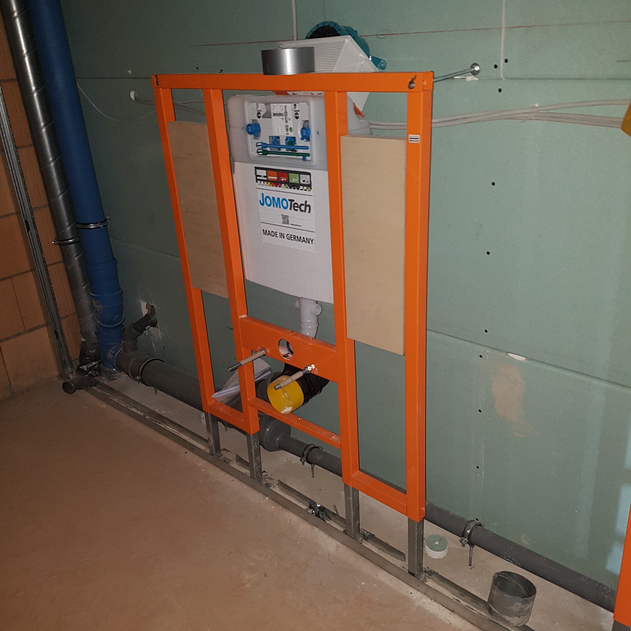 Pomarańczowa rama z zamontowanym stelażem podtynkowym JOMOTech, widoczne rury kanalizacyjne i fragment ściany z płyt karton-gips, etap prac instalacyjnych.