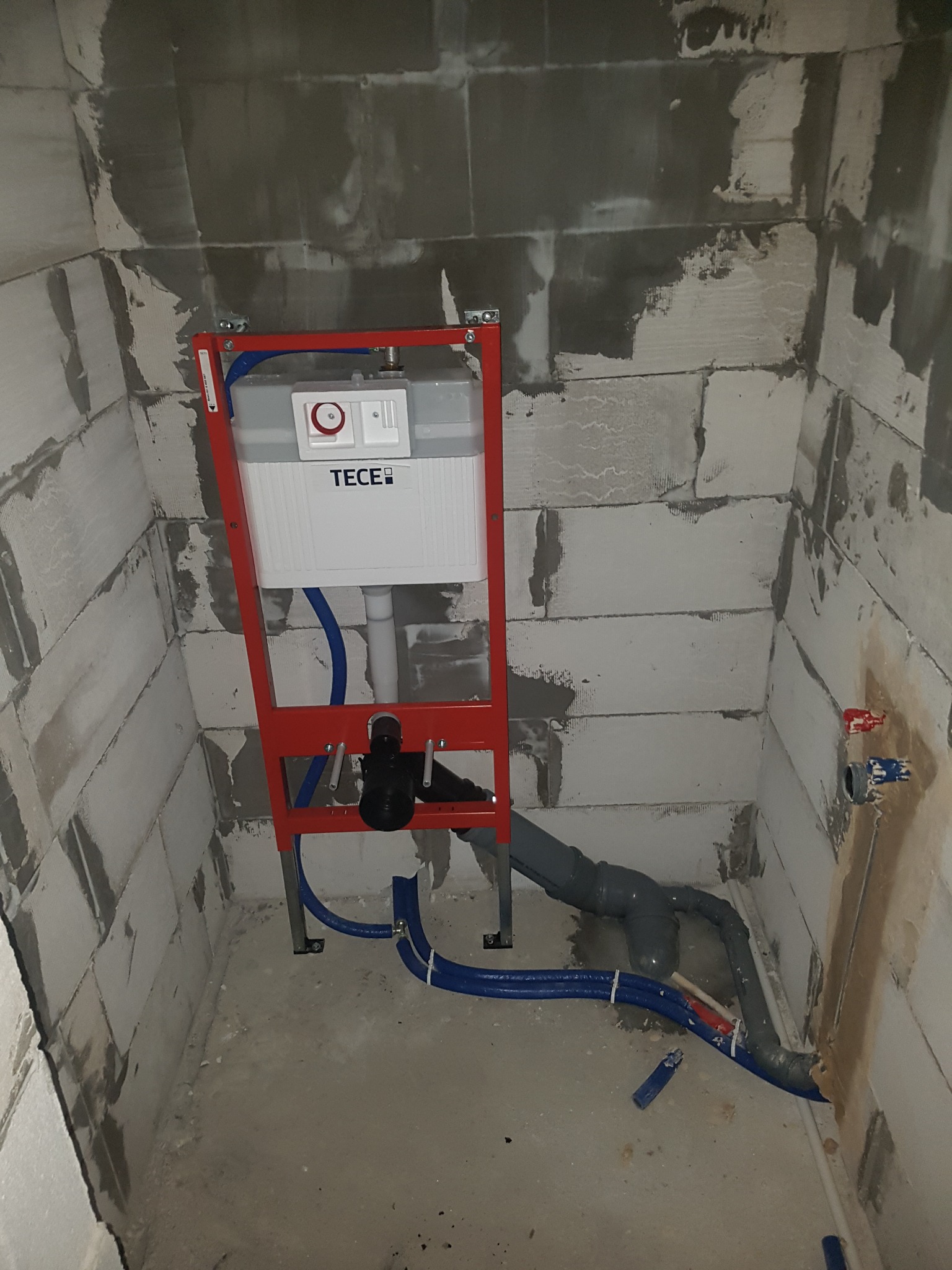 Stelaż podtynkowy WC marki TECE z częściowo podłączonymi rurami, widoczne przyłącza wody ciepłej i zimnej na surowej ścianie z betonu komórkowego.