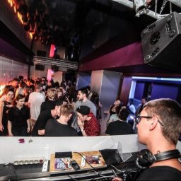 Łukasz Baumgart - DJ w trakcie występu w klubie, widoczny tłum ludzi, oświetlenie dyskotekowe i sprzęt nagłośnieniowy. DJ ma na głowie słuchawki i skupia się na miksowaniu muzyki.