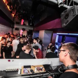 DJ w trakcie występu w klubie, widoczny tłum ludzi, oświetlenie dyskotekowe i sprzęt nagłośnieniowy. DJ ma na głowie słuchawki i skupia się na miksowaniu muzyki.