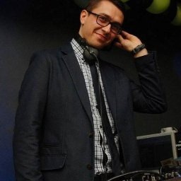 Łukasz Baumgart - Uśmiechnięty DJ w marynarce i słuchawkach, z ręką przy uchu, stoi za konsoletą na tle czarnych i zielonych balonów, gotowy do występu.