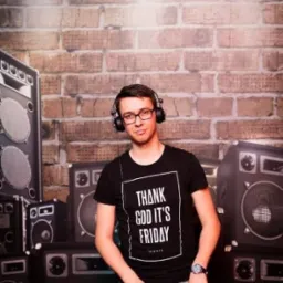 DJ w czarnej koszulce z napisem 'Thank God It's Friday' miksuje muzykę na tle ceglanej ściany i kolumn głośnikowych.
