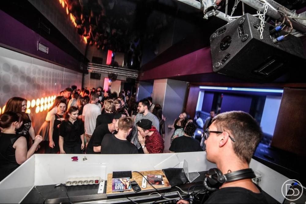 DJ w trakcie występu w klubie, widoczny tłum ludzi, oświetlenie dyskotekowe i sprzęt nagłośnieniowy. DJ ma na głowie słuchawki i skupia się na miksowaniu muzyki.