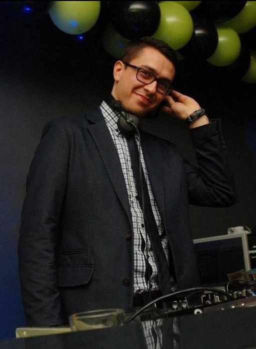 Uśmiechnięty DJ w marynarce i słuchawkach, z ręką przy uchu, stoi za konsoletą na tle czarnych i zielonych balonów, gotowy do występu.