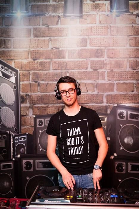 DJ w czarnej koszulce z napisem 'Thank God It's Friday' miksuje muzykę na tle ceglanej ściany i kolumn głośnikowych.