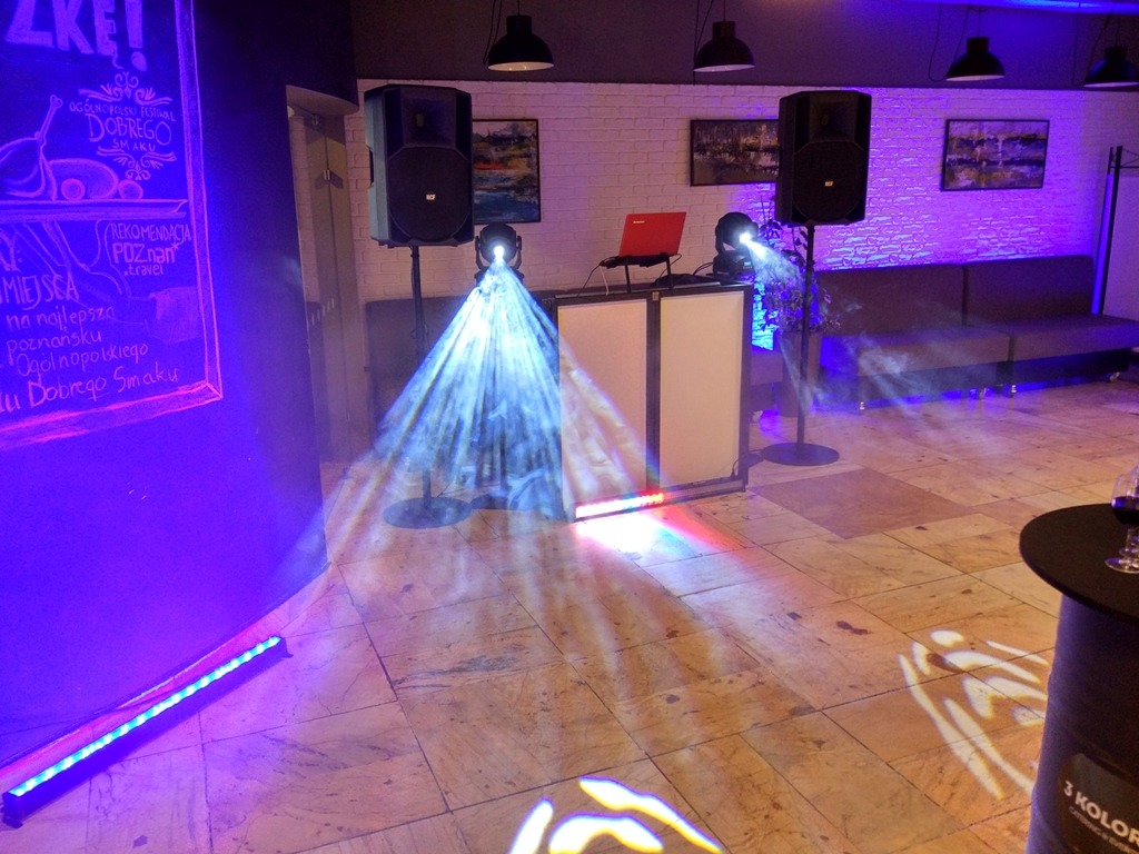 Sala z nagłośnieniem i oświetleniem eventowym: kolumny głośnikowe, konsoleta DJ-ska z laptopem, reflektory tworzące snopy światła, podświetlona ściana z napisem.