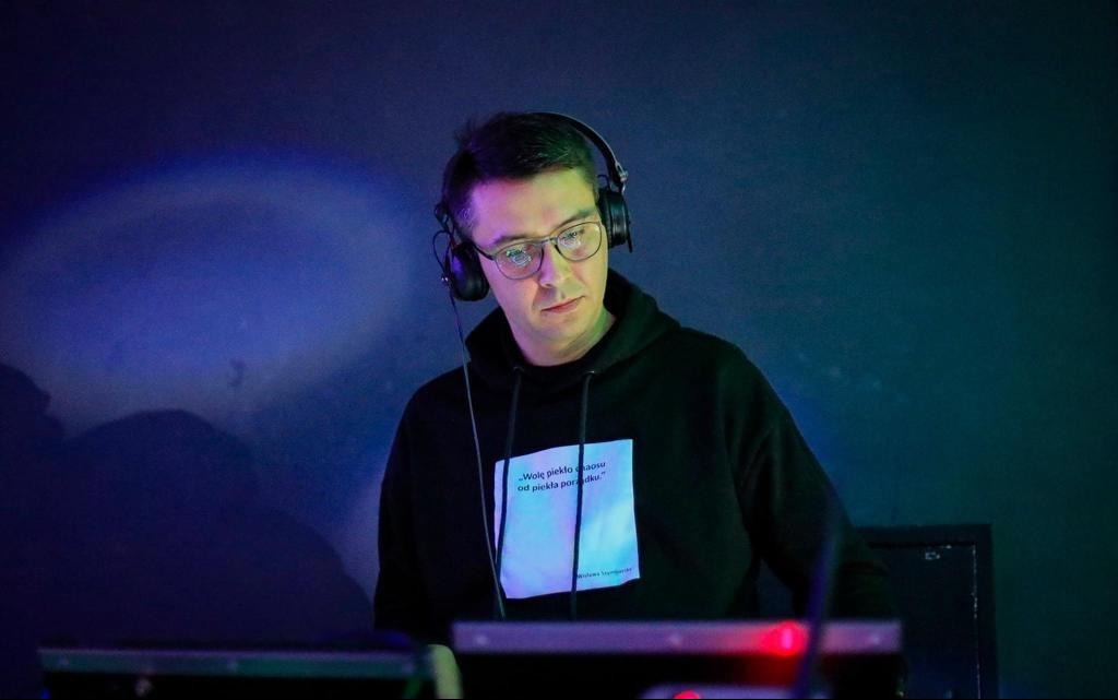 DJ w słuchawkach, skupiony przy konsolecie, w tle ciemne oświetlenie sceniczne. Na bluzie widoczny cytat.