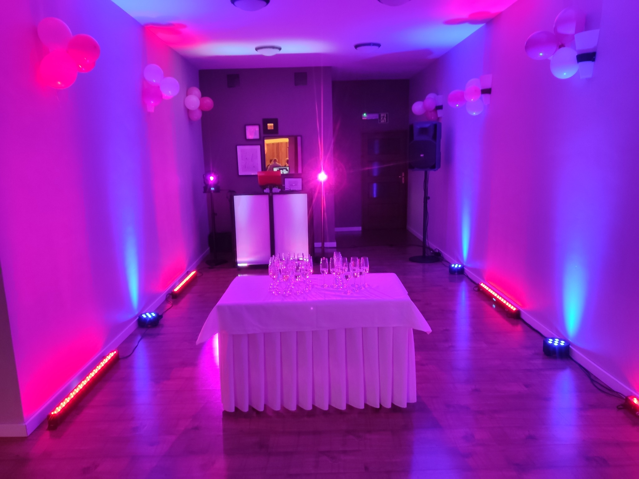 Szykowna sala bankietowa z dekoracją balonową, stołem z kieliszkami i profesjonalnym oświetleniem w odcieniach różu i fioletu, gotowa na imprezę.