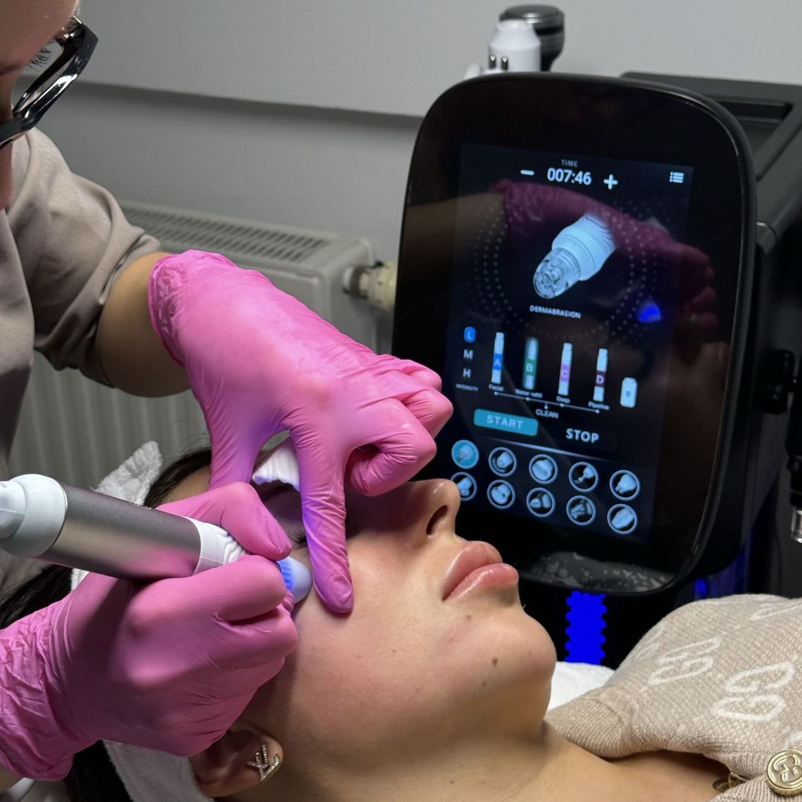 Zabieg hydradermabrazji na twarzy klientki wykonywany przez kosmetyczkę w różowych rękawiczkach. Widoczny ekran urządzenia z ustawieniami.