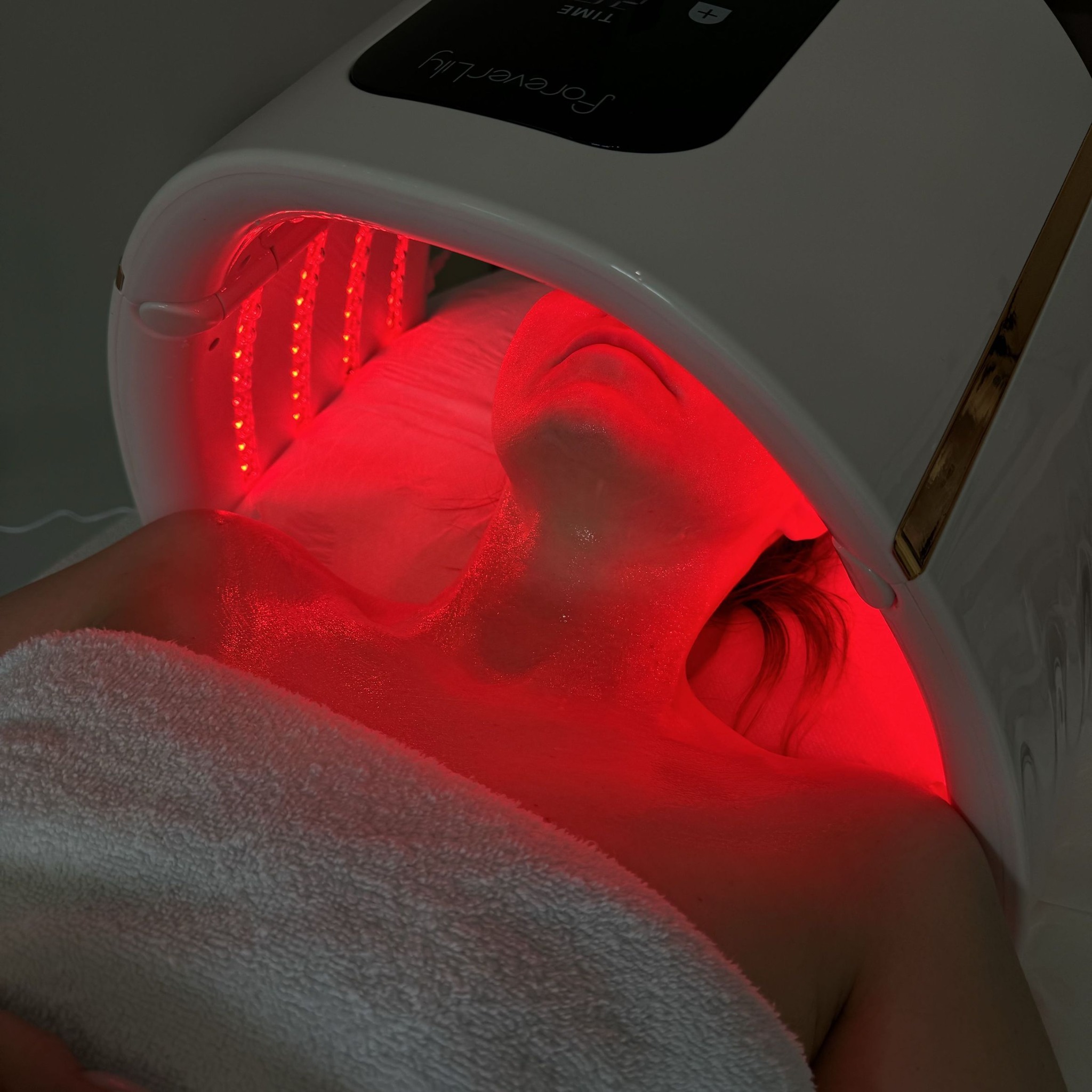 Klientka poddawana zabiegowi na szyję i dekolt z użyciem lampy LED Foreverlily w salonie. Czerwone światło oświetla skórę, która wydaje się być nawilżona.