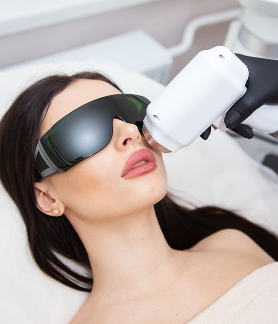 Kobieta w okularach ochronnych poddawana zabiegowi laserowemu na twarzy. Widoczny aplikator i dłoń w czarnej rękawiczce.