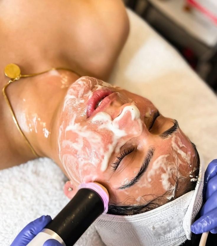 Klientka z pianką na twarzy poddawana zabiegowi upiększającemu z użyciem specjalistycznego urządzenia, trzymanego przez osobę w niebieskich rękawiczkach.