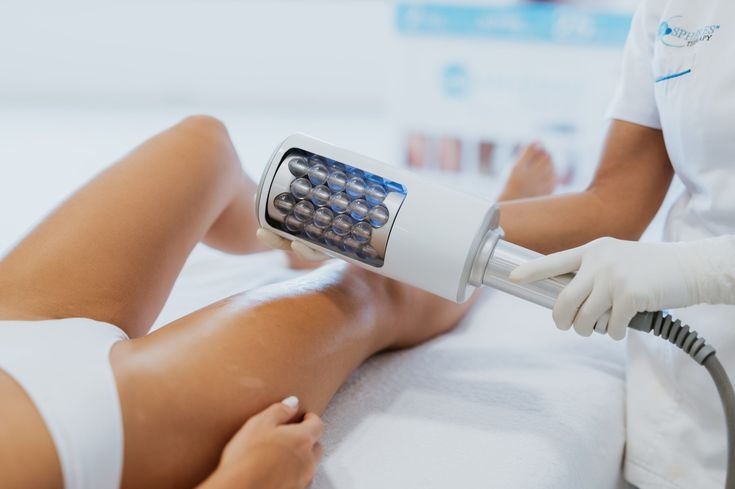 Zabieg na nogi urządzeniem z kulkami w salonie kosmetycznym. Widoczne dłonie w rękawiczkach i fragment białego uniformu osoby wykonującej zabieg.