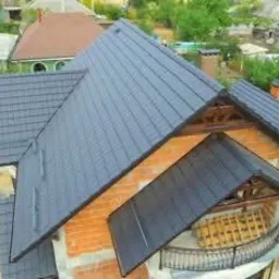 Nowy, ciemnoszary dach z blachodachówki na budynku z surowej cegły, widok z góry. W tle zabudowa jednorodzinna i zieleń drzew.