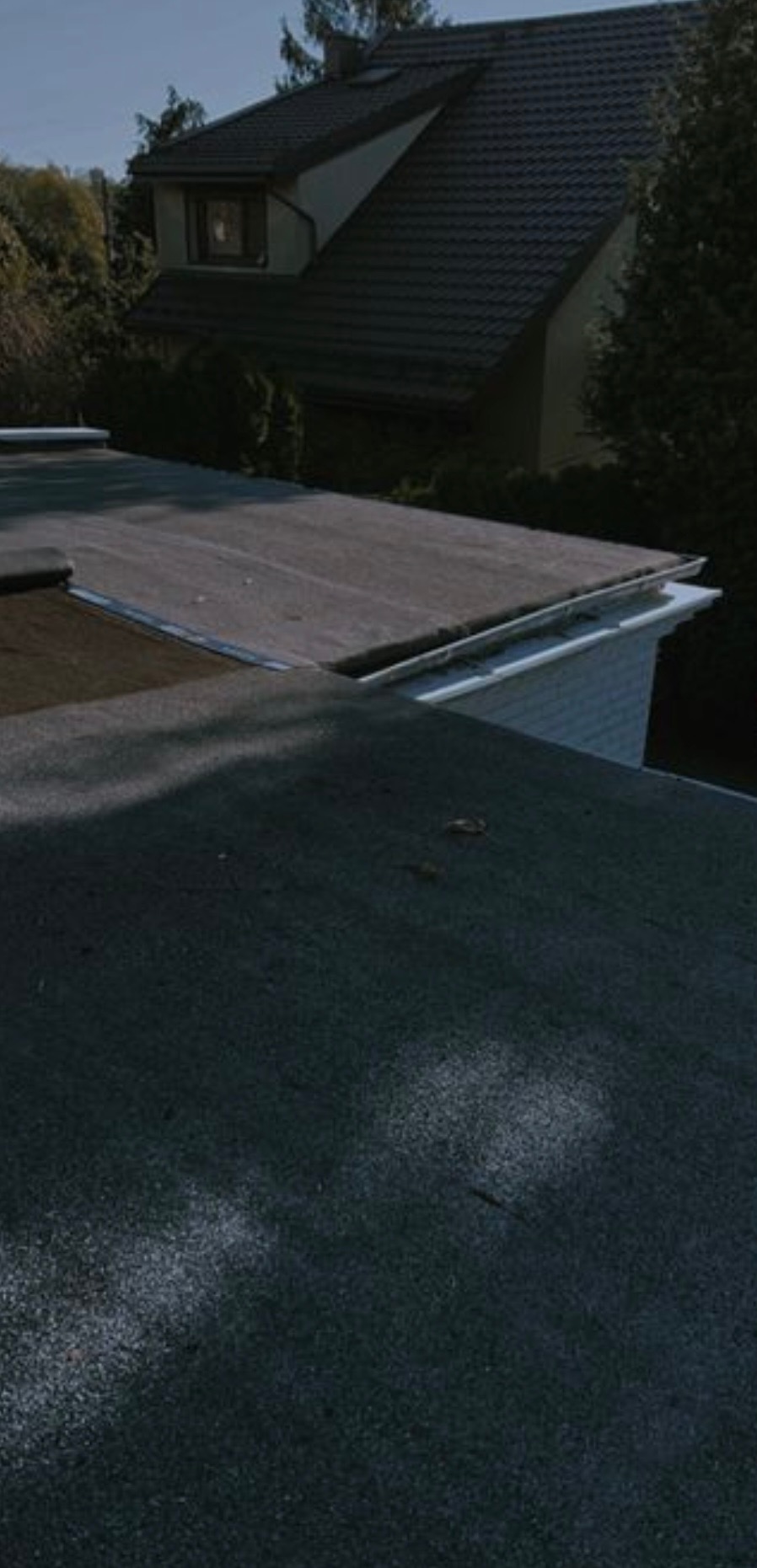 Widok z góry na płaski dach pokryty papą, z widoczną rynną i fragmentem budynku w tle, otoczonego zielenią. Dach w odcieniach szarości, rynna biała.