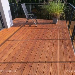 Adam Tomczyk - Nowoczesny taras z drewna kompozytowego, z minimalistycznym krzesłem i donicą z trawą ozdobną. Szklane balustrady podkreślają elegancki design.