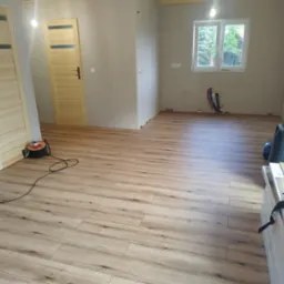 Jasne panele podłogowe ułożone w nowym pokoju, widoczne narzędzia i paczki paneli. Ściany przygotowane do malowania, w tle okno z widokiem na zieleń.