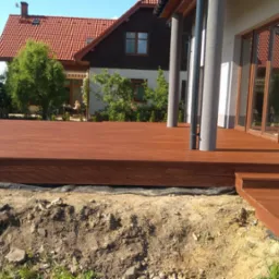 Rozległy taras z drewna o ciepłym, brązowym odcieniu, z niskimi schodami, idealnie komponujący się z architekturą domu i zielenią ogrodu w słoneczny dzień.