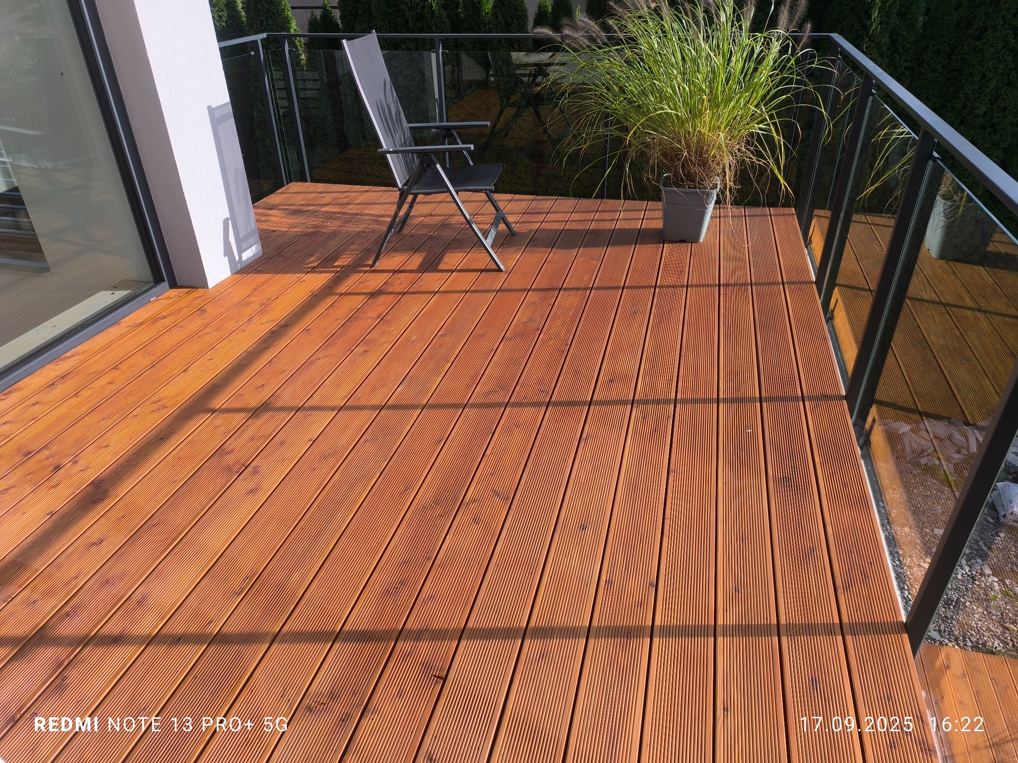 Nowoczesny taras z drewna kompozytowego, z minimalistycznym krzesłem i donicą z trawą ozdobną. Szklane balustrady podkreślają elegancki design.