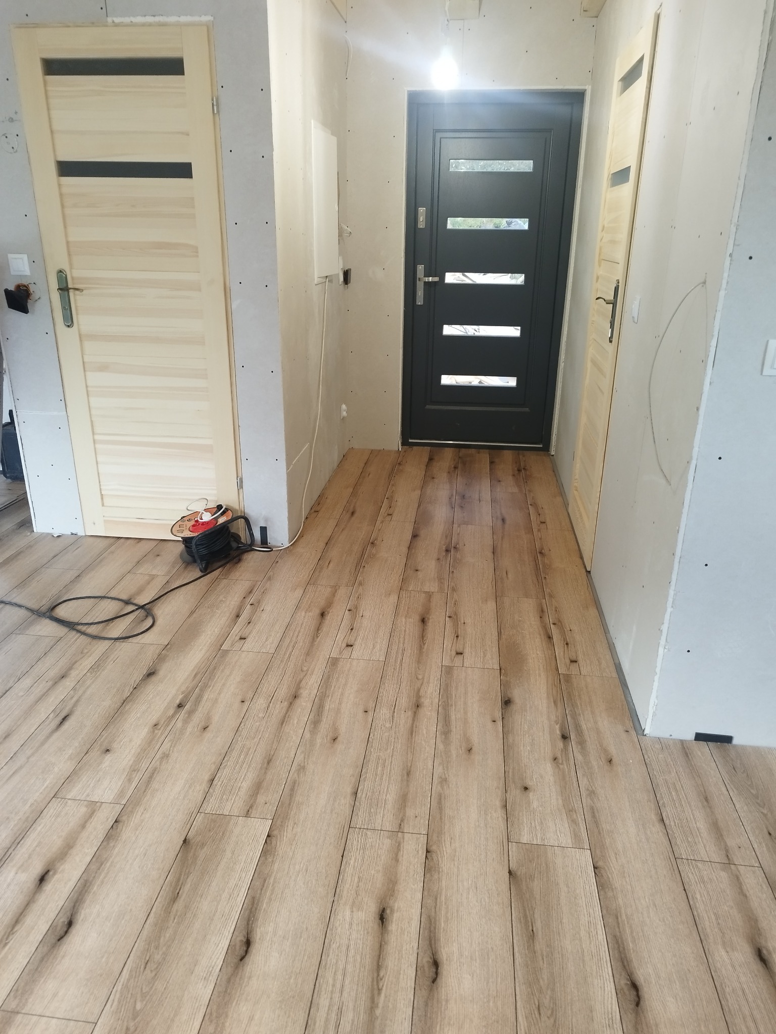 Jasne panele podłogowe ułożone w korytarzu z dwiema parami drzwi i surowymi ścianami, widoczny zwinięty przedłużacz elektryczny.