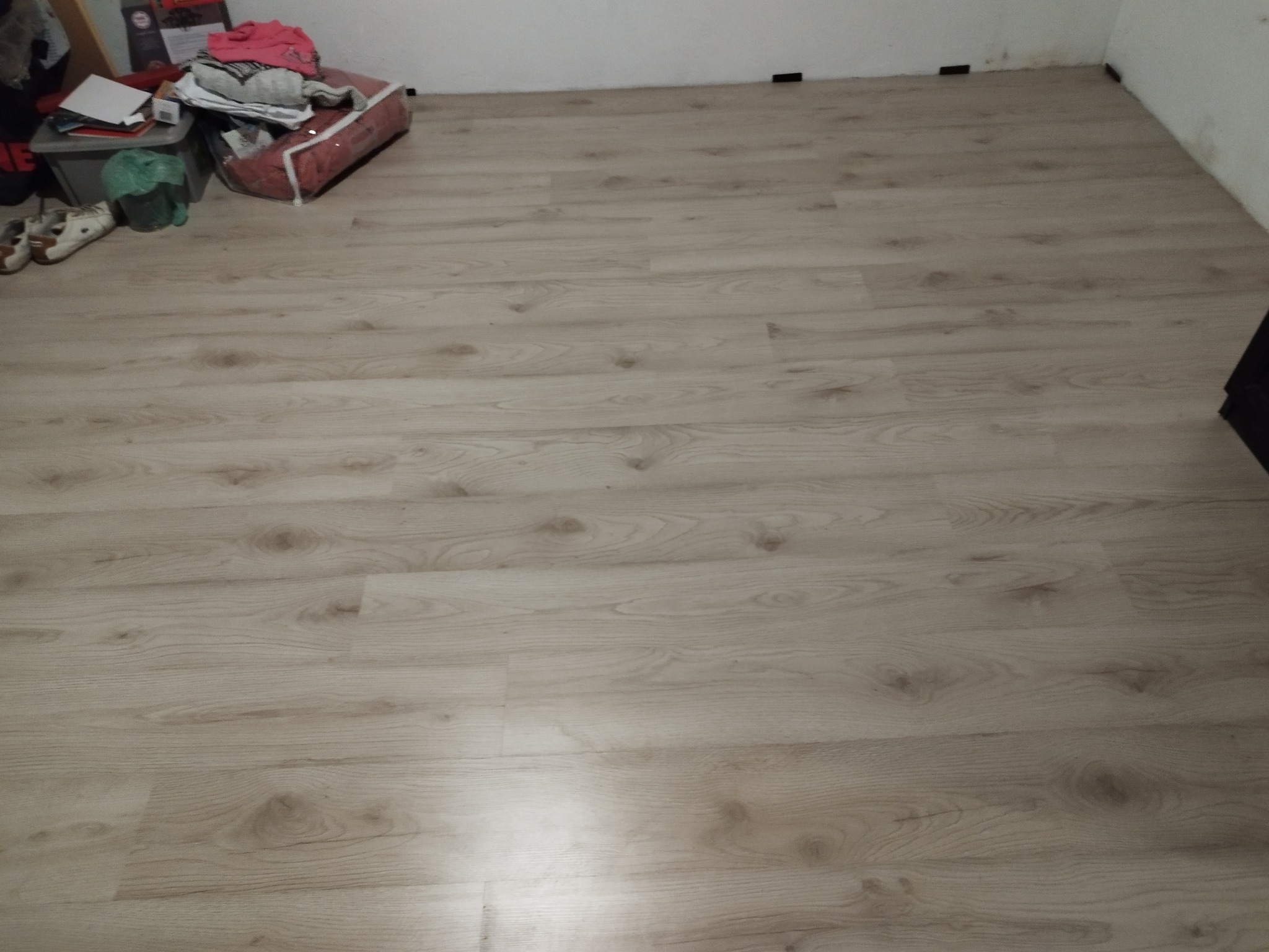 Jasne panele podłogowe z widocznym wzorem drewna, ułożone w pomieszczeniu. W rogu widoczne przedmioty zakrywające fragment podłogi. Widok z góry.