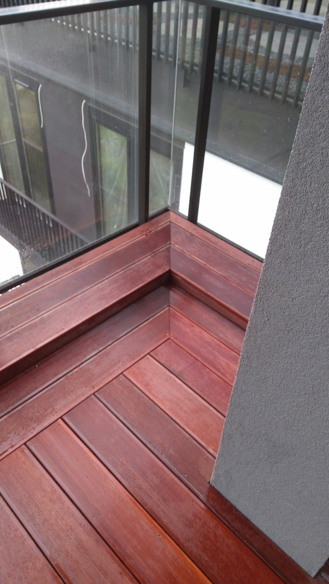 Czerwono-brązowy taras drewniany z widocznymi deskami ułożonymi w jodełkę, otoczony szklaną balustradą i fragmentem ściany o szarej, chropowatej fakturze.