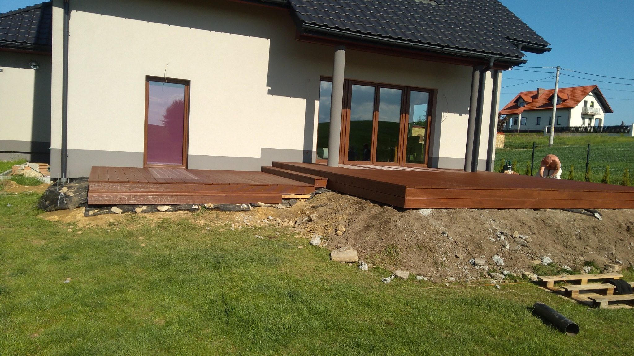 Nowoczesny taras drewniany z brązowych desek przy domu z ciemną dachówką. Widoczne schody tarasowe i przygotowane podłoże. W tle dom sąsiada i pracownik.