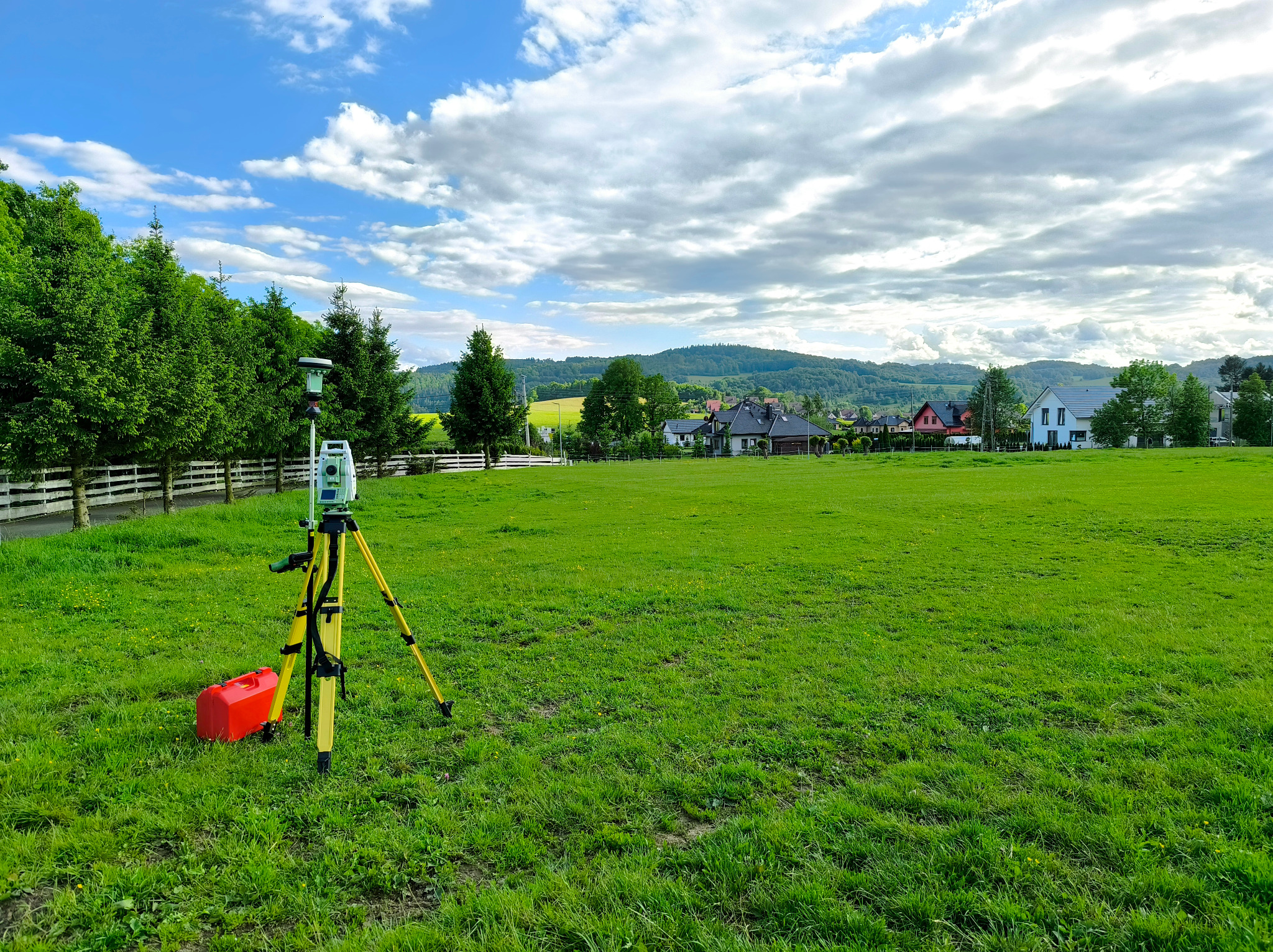 Tachimetr na statywie z czerwoną skrzynką narzędziową na zielonym polu, w tle drzewa, domy i wzgórza pod błękitnym niebem z chmurami. Sprzęt pomiarowy.