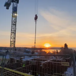 Wczesny etap budowy domu murowanego zimą, z widocznym żurawiem budowlanym i zbrojeniami fundamentów na tle wschodzącego słońca nad zamarzniętym jeziorem.