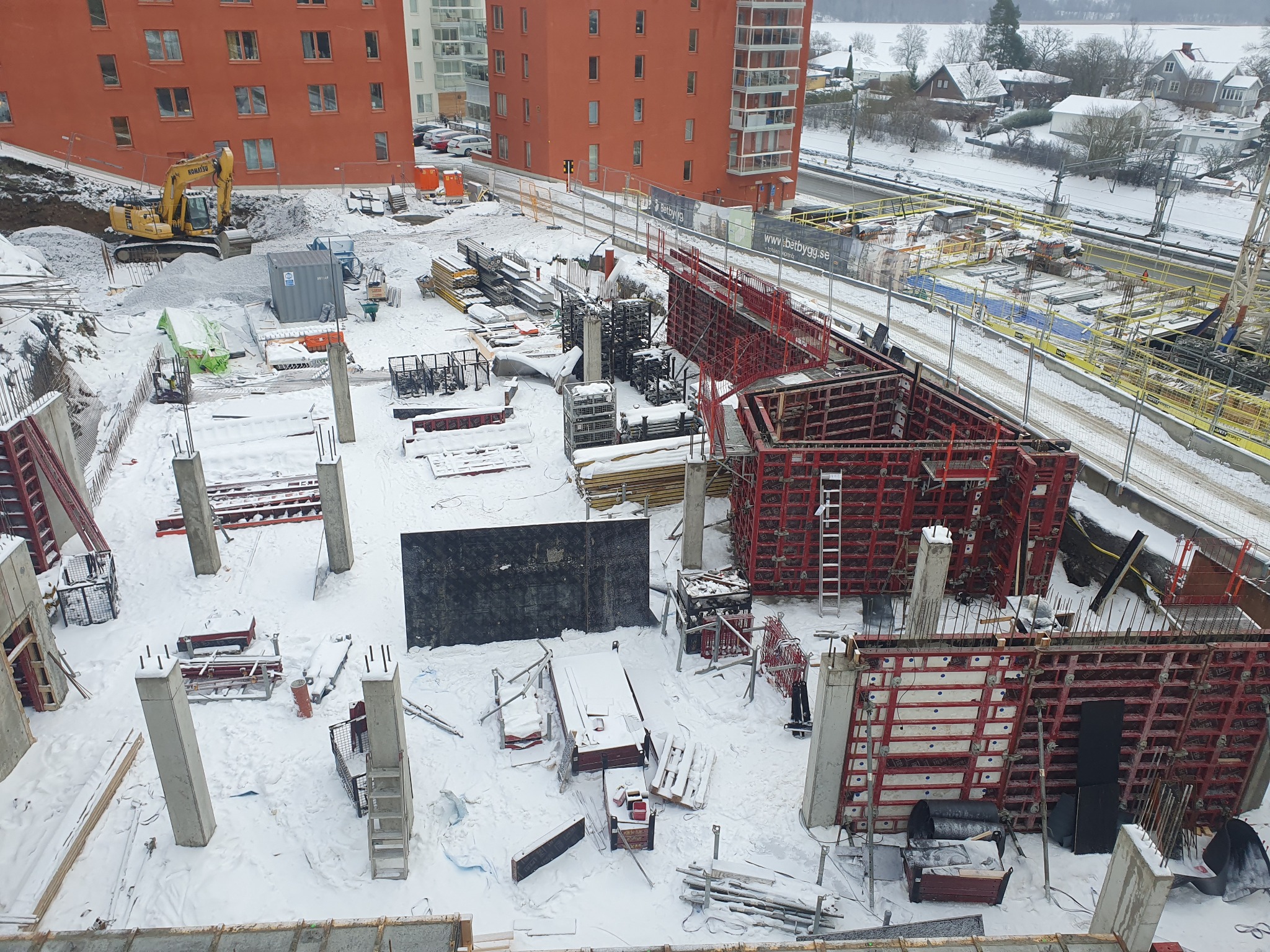 Zimowy plac budowy z widokiem na fundamenty, kolumny i szalunki w czerwieni, z żółtą koparką w tle i zasypanym śniegiem otoczeniem.