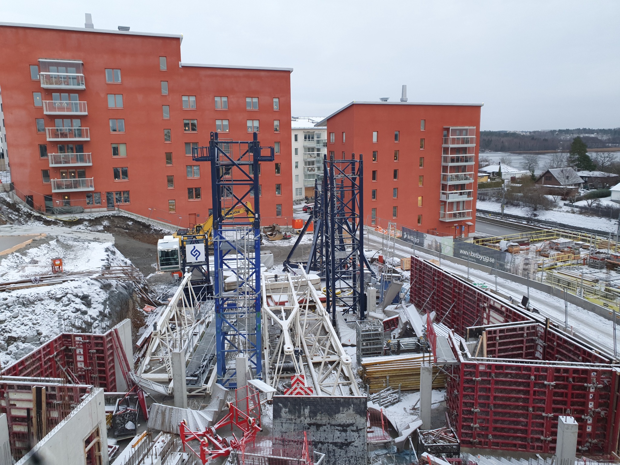 Zimowy plac budowy z czerwonymi budynkami w tle. Widoczne szalunki, żurawie i elementy konstrukcyjne pokryte śniegiem. Chłodna kolorystyka.