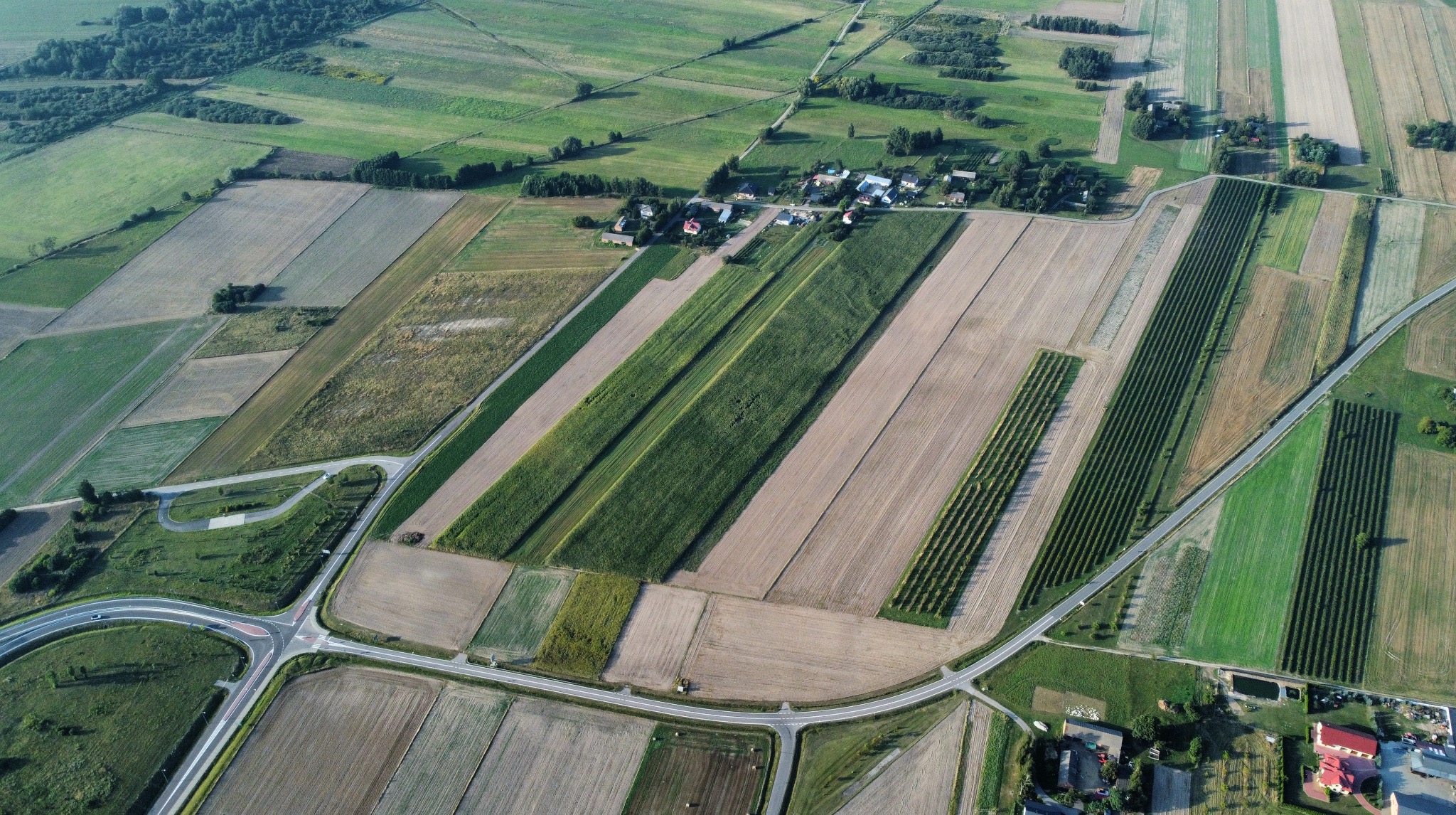 Panoramiczny widok z lotu ptaka na mozaikę pól uprawnych, przecinanych drogami, z widoczną małą wioską w centralnej części kadru, w słoneczny dzień.