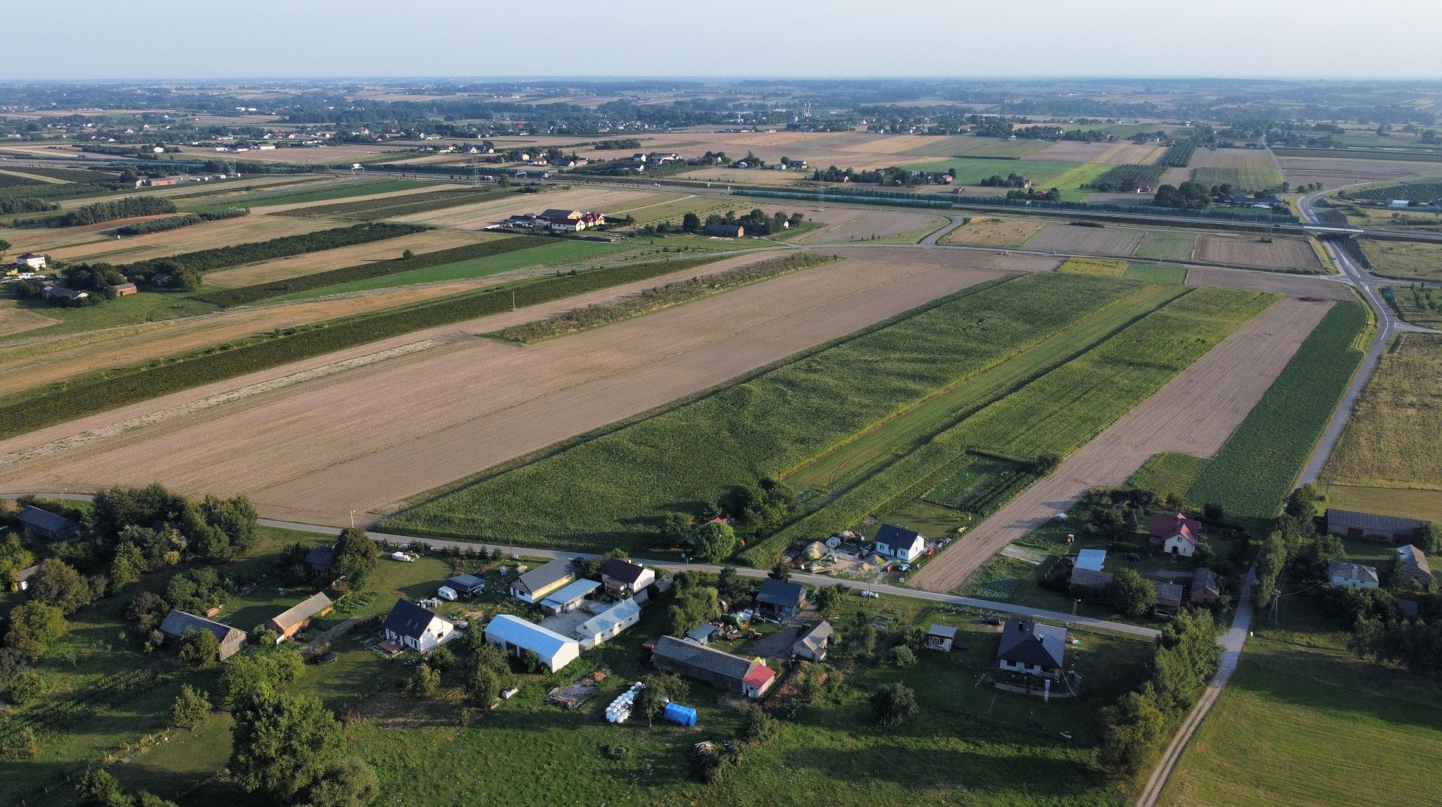 Szeroki kadr z lotu ptaka ukazuje mozaikę pól uprawnych, przeplatanych drogami i zabudowaniami wiejskimi. W dole widoczne domy z czerwonymi dachami.
