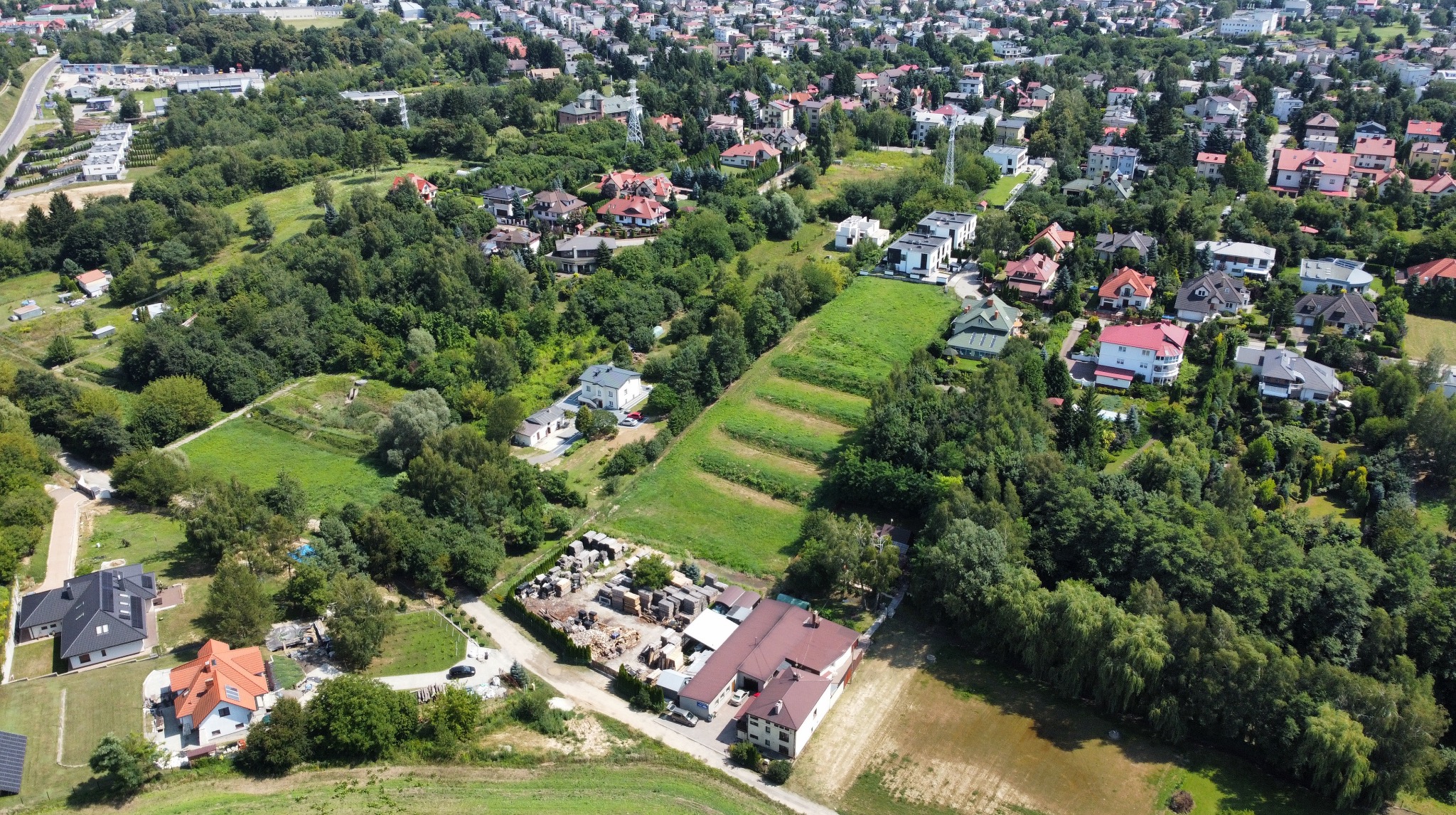 Panoramiczny widok z lotu ptaka na osiedle domów jednorodzinnych otoczonych zielenią, w tle widoczny fragment miasta.