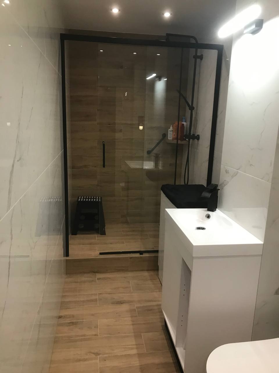 Nowoczesna łazienka z kabiną prysznicową typu walk-in, wykończona płytkami imitującymi drewno i marmur, z czarnymi akcentami i białymi meblami.