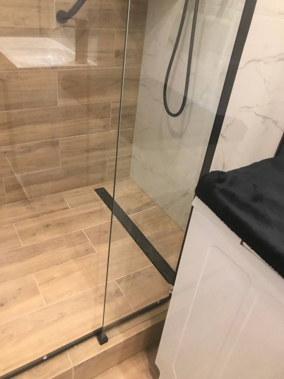 Nowoczesna kabina prysznicowa z czarnymi profilami i odpływem liniowym. Beżowe płytki imitujące drewno na podłodze i marmurowe na ścianach.
