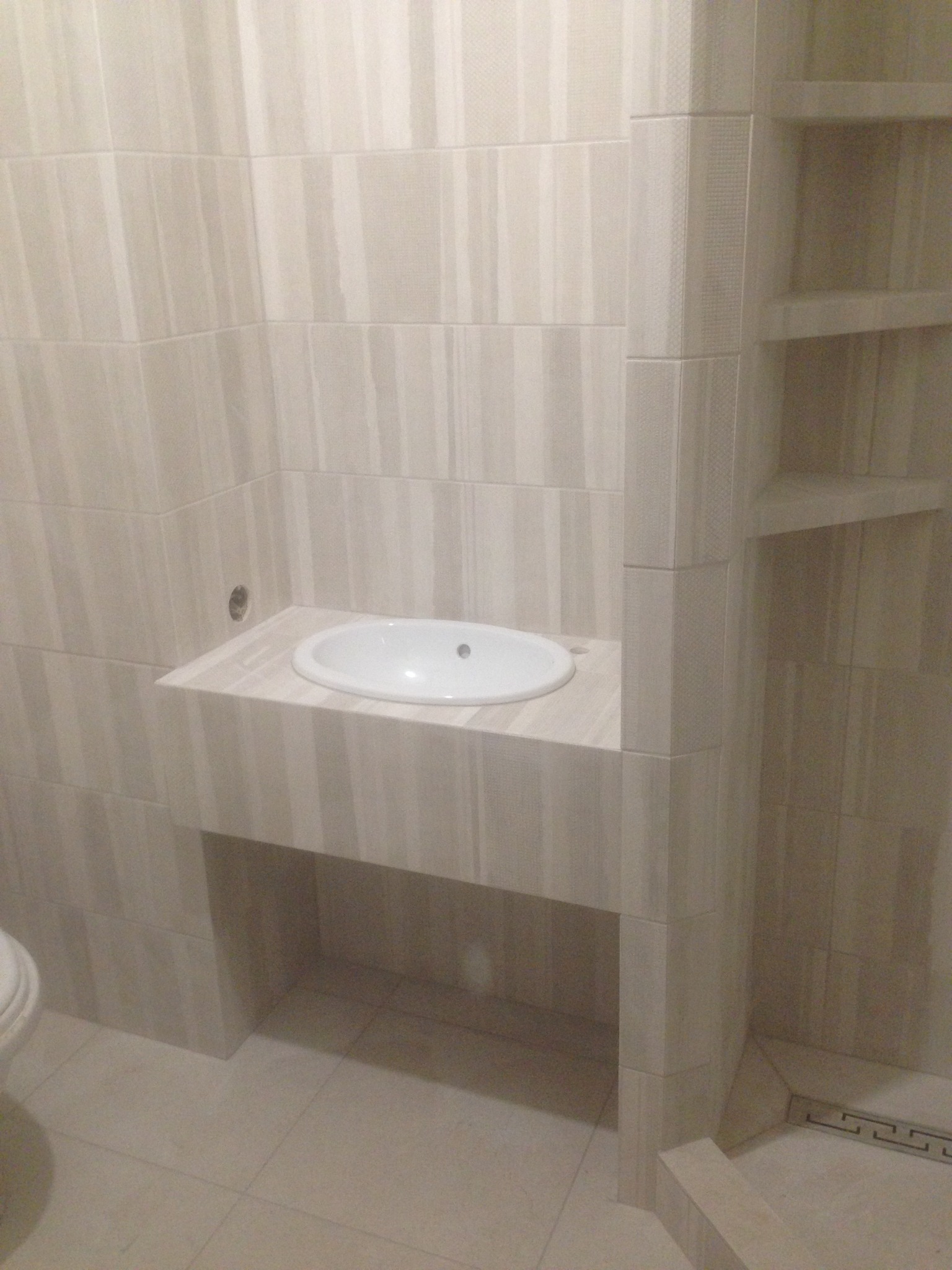 Wykończona łazienka z umywalką i półkami w beżowych płytkach. Widoczny fragment toalety i odpływ liniowy w podłodze. Ściany w pionowe pasy.