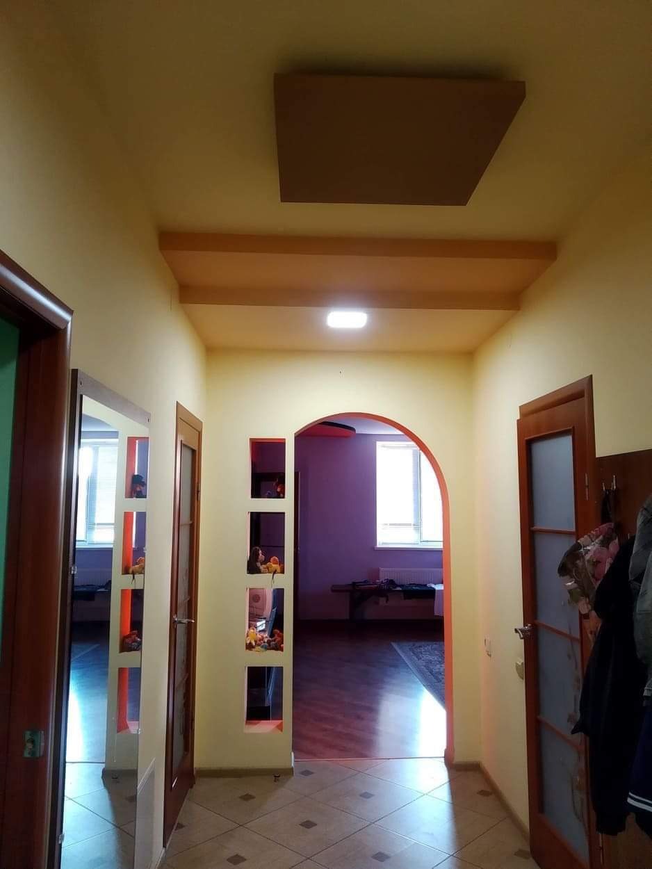 Korytarz po remoncie w Warszawie: żółte ściany, drewniane elementy sufitu i drzwi, łukowate przejście do pokoju, wnęki w ścianie z dekoracjami.