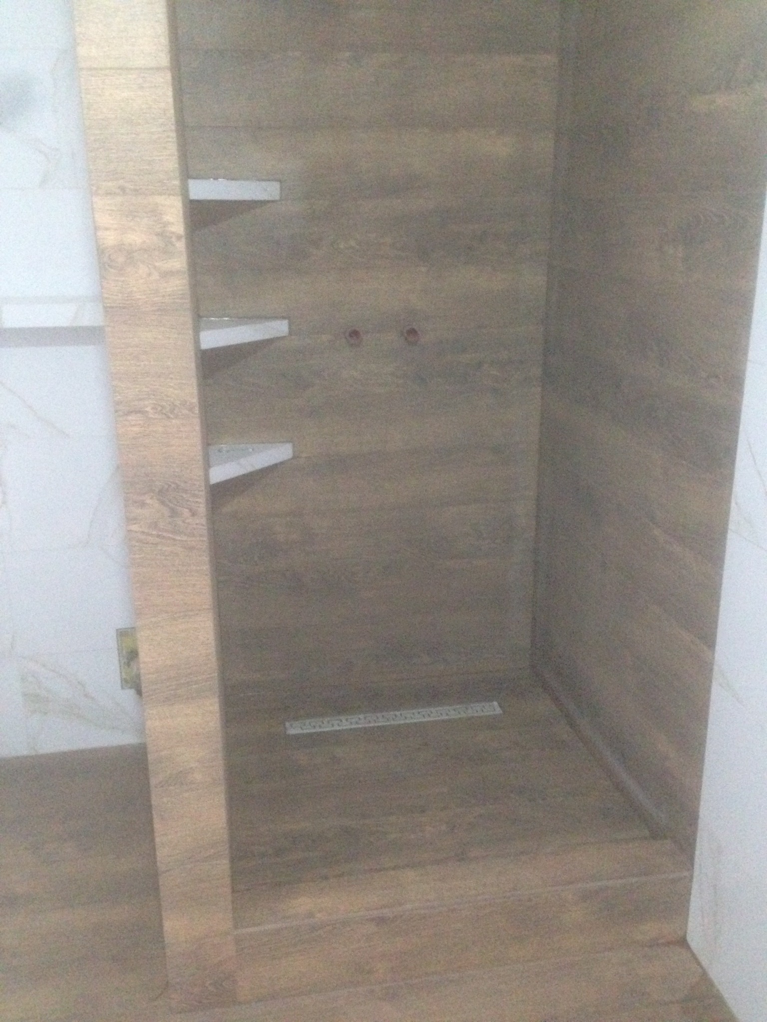 Wykończona kabina prysznicowa z drewnopodobnymi płytkami, widoczne półki i odpływ liniowy. Minimalistyczny design, ciepłe odcienie brązu.