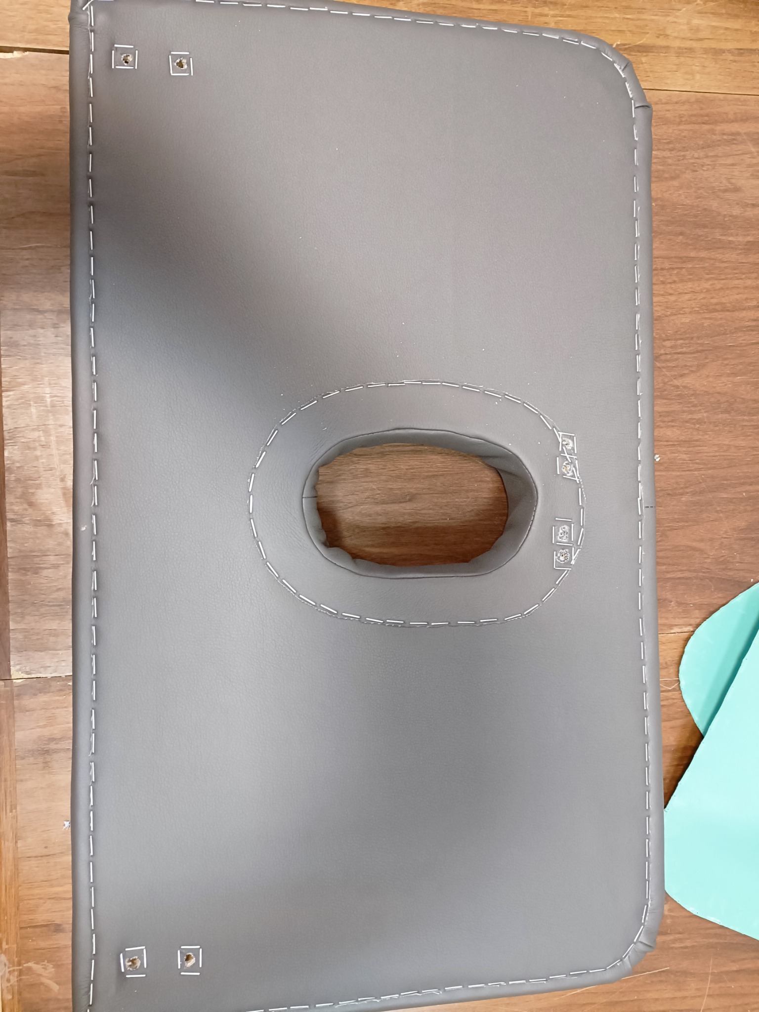 Szary pokrowiec z otworem na twarz, obszyty białą nicią, leżący na drewnianej powierzchni. Widoczne zszywki i fragment turkusowego materiału.