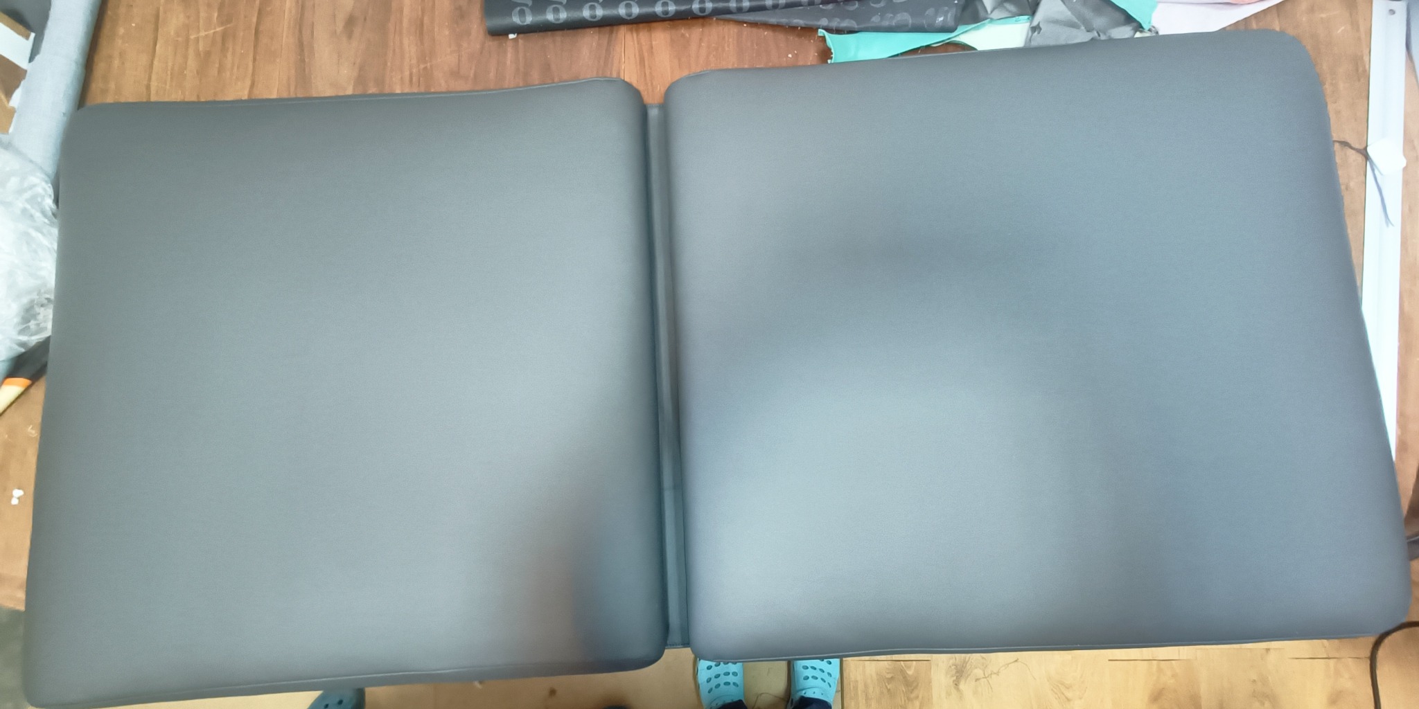 Szary pokrowiec na dwuczęściowy materac, leżący na drewnianej podłodze, widok z góry. Detal szwu i zaokrąglonych krawędzi. W tle materiały i fragment obuwia.