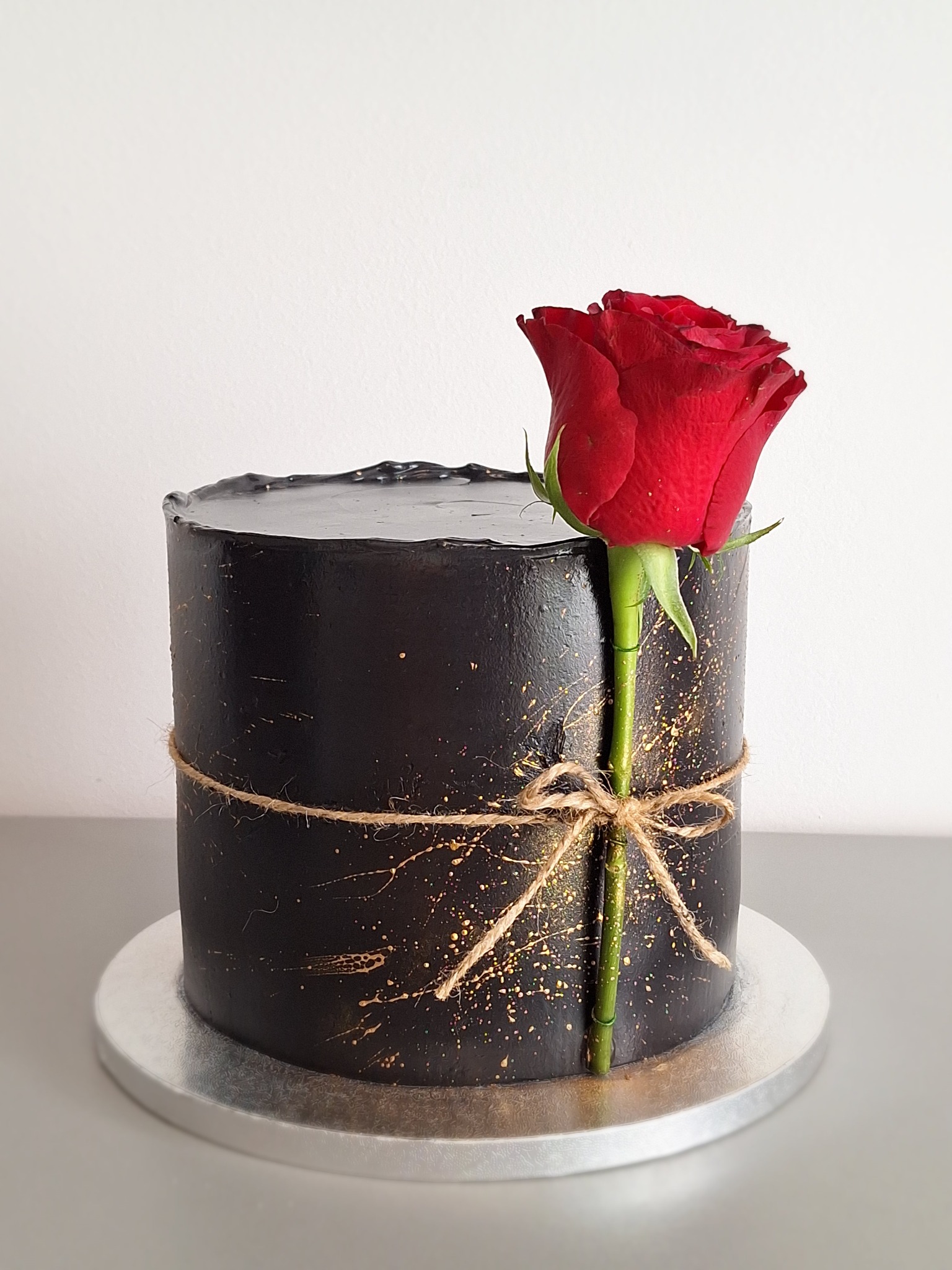 Czarny tort z czerwoną różą, owinięty jutowym sznurkiem i ozdobiony złotymi plamkami na srebrnej podstawie. Minimalistyczny styl.