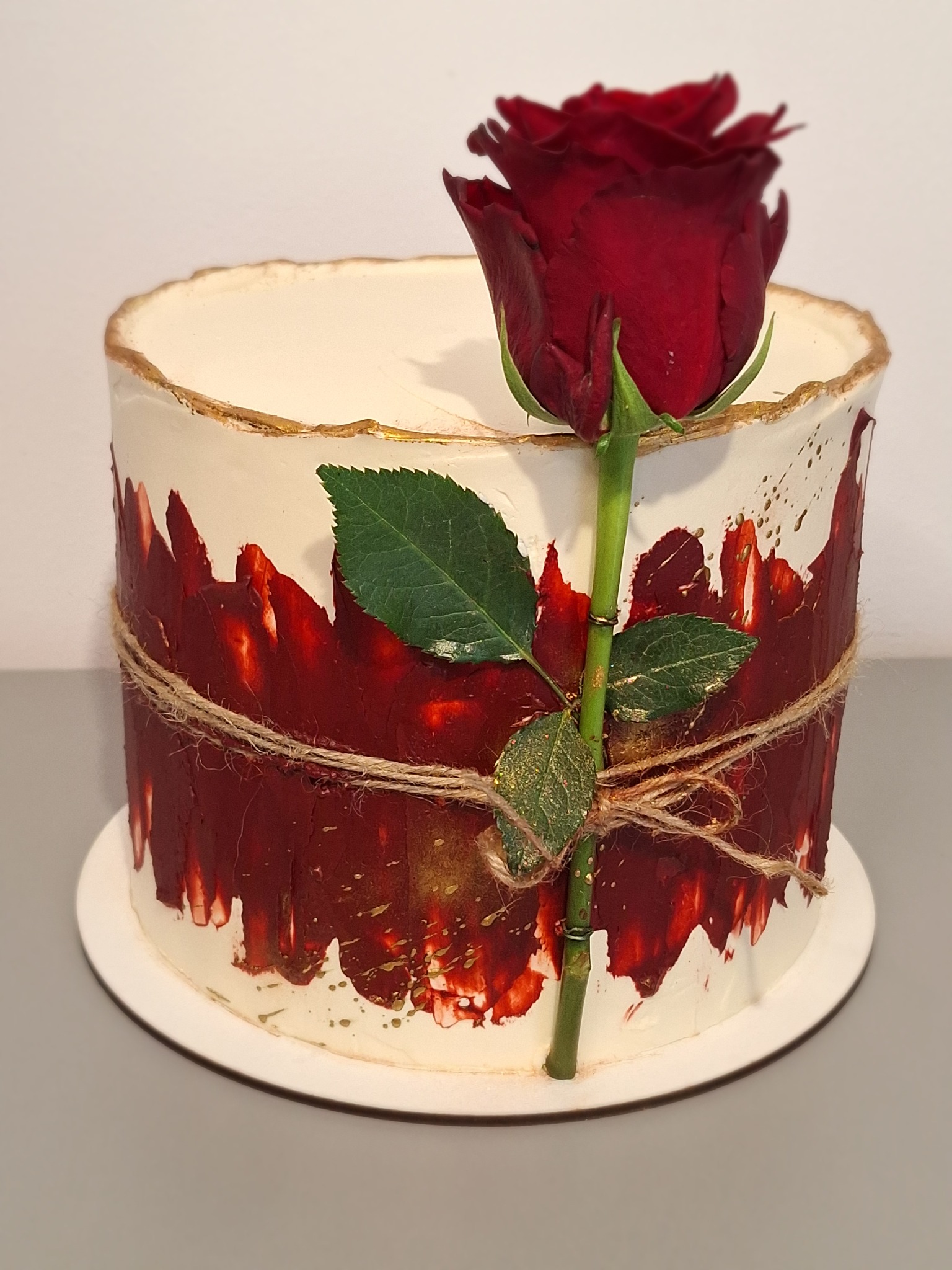 Tort artystyczny z kremem, zdobiony czerwonymi plamami, złotem i sznurkiem jutowym, zwieńczony pojedynczą, ciemnoczerwoną różą. Elegancki i unikalny.
