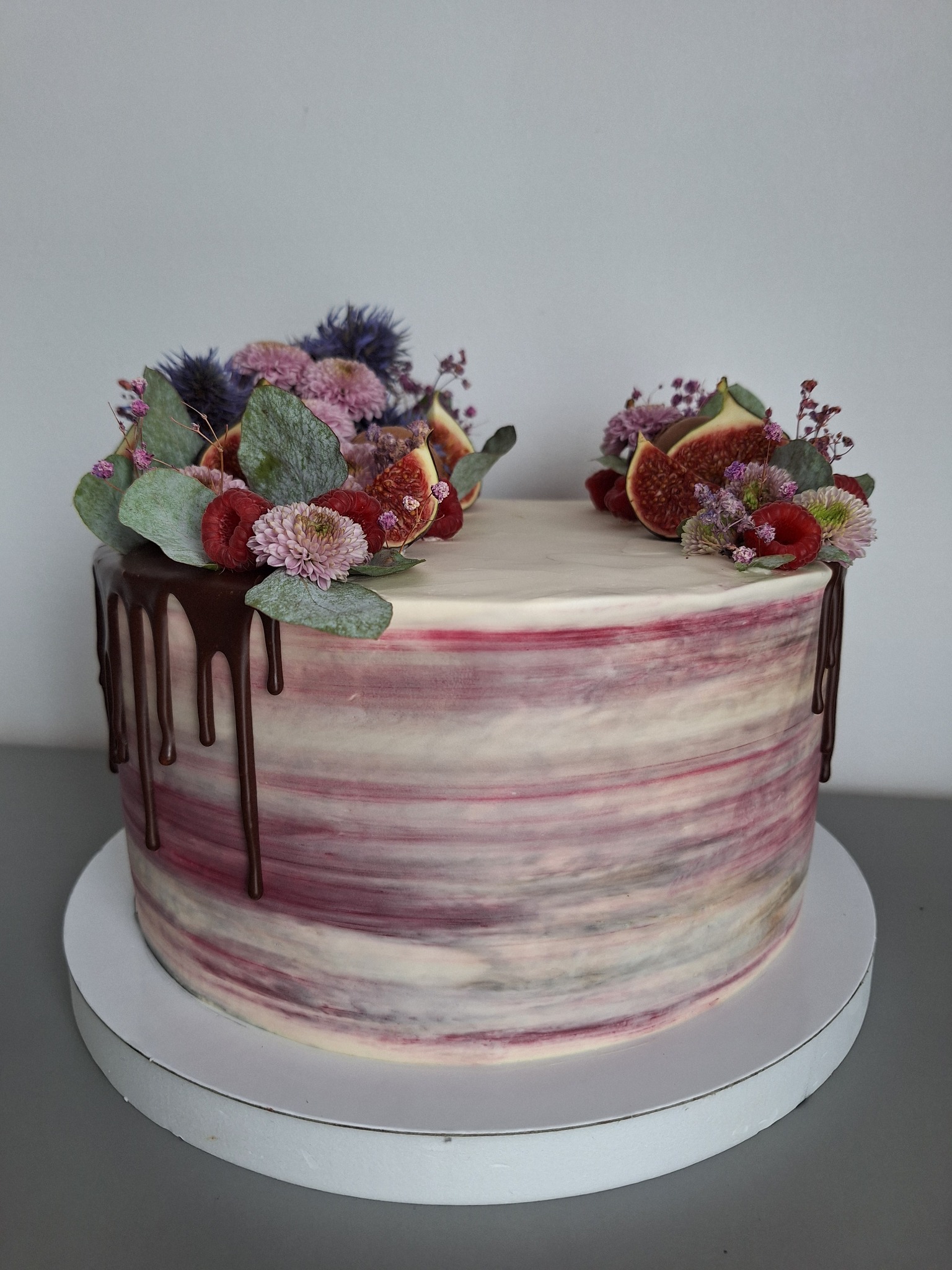 Tort z kremem w odcieniach różu i bieli, udekorowany świeżymi figami, malinami, zielonymi liśćmi i fioletowymi kwiatami, z polewą czekoladową spływającą po bokach.