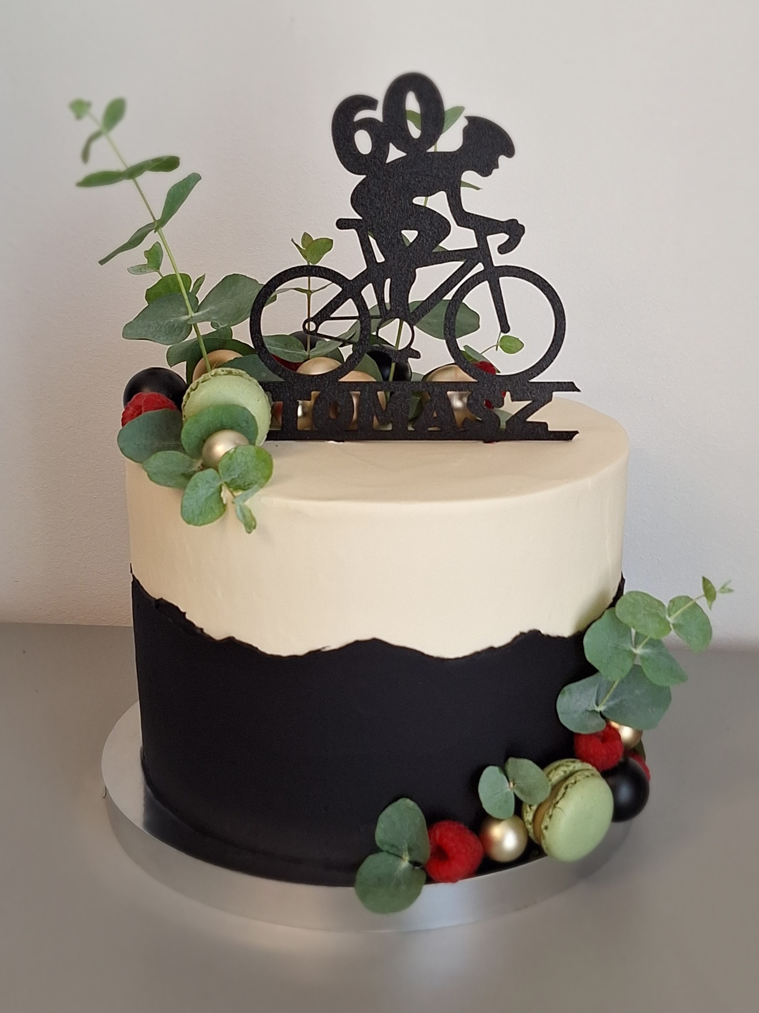 Tort dwukolorowy z topperem rowerzysty i napisem 'Tomasz', dekorowany makaronikami, malinami i eukaliptusem. Minimalistyczny styl, idealny na 60. urodziny.