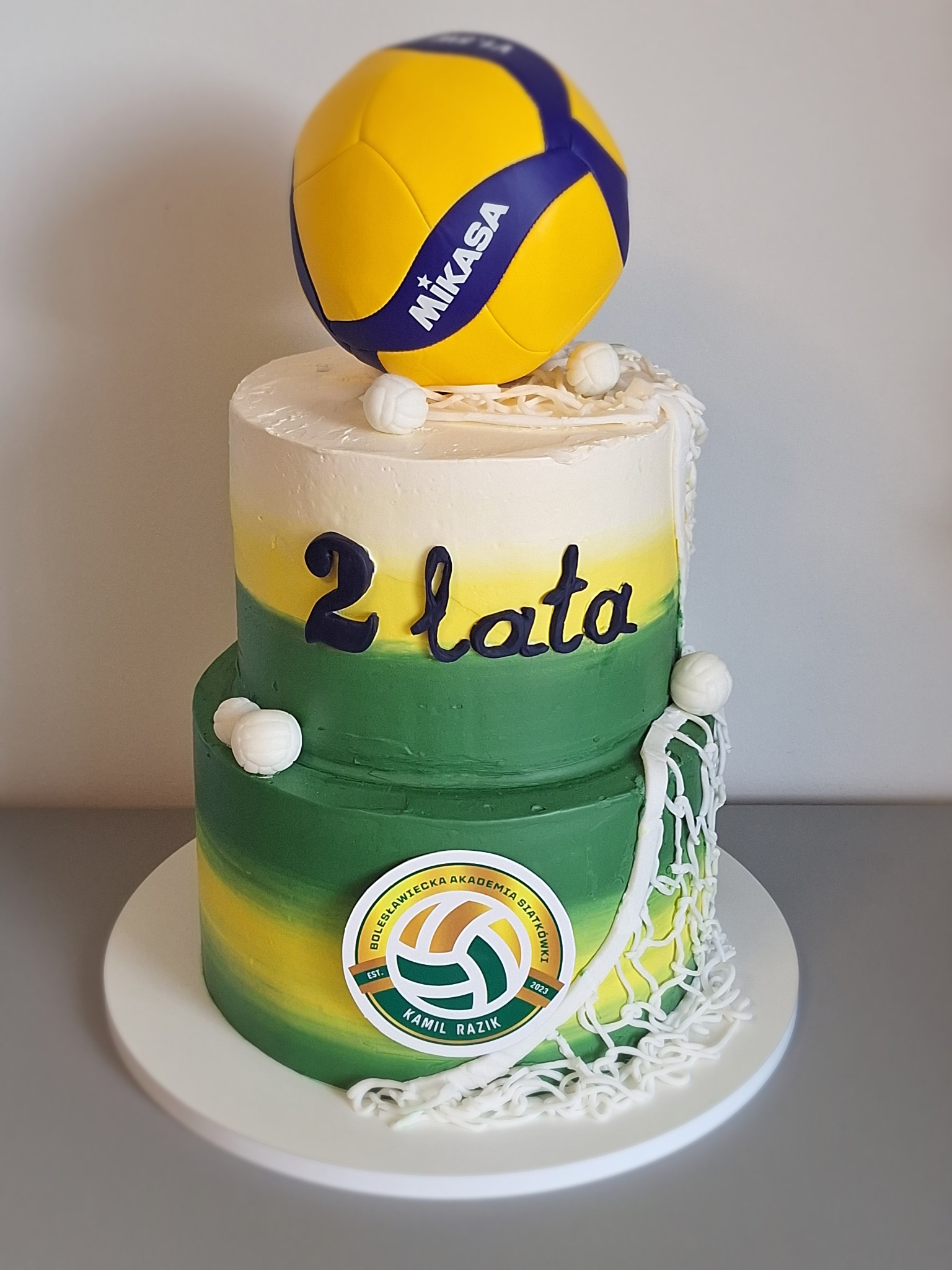 Dwupiętrowy tort w barwach klubu siatkarskiego, z piłką Mikasa na wierzchu, dekorowany logiem klubu, napisem '2 lata' i imitacją siatki. Całość utrzymana w tonacji zieleni, żółci i bieli.